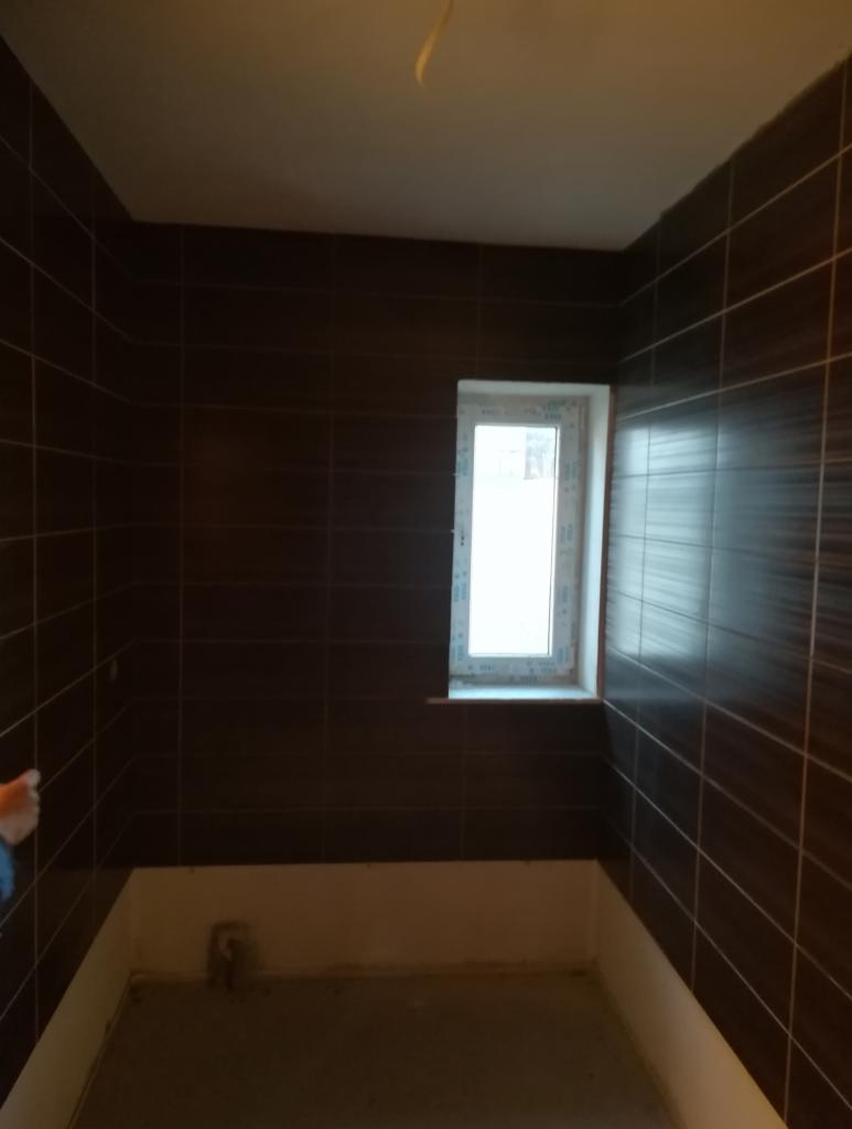 Brązowe, prostokątne płytki ułożone na ścianach w łazience, widoczne białe okno i przygotowane podłoże pod wannę lub prysznic.