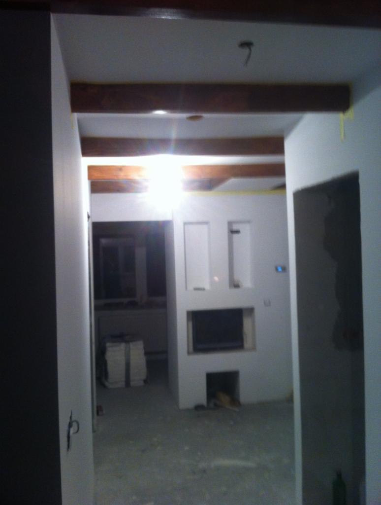 Wnętrze w trakcie prac, widoczne surowe ściany, drewniane belki stropowe, przygotowane miejsce na kominek oraz materiały budowlane w tle.