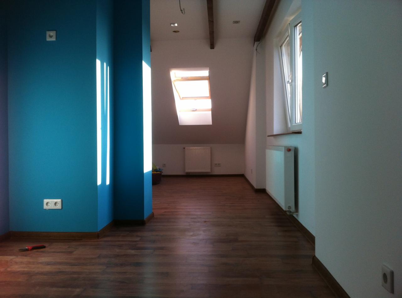 Poddasze po remoncie: turkusowa ściana z promieniami słońca kontrastuje z drewnianą podłogą i białymi ścianami, widoczne okno dachowe i grzejniki.