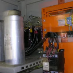 Wnętrze szafy elektrycznej z kondensatorami mocy, okablowaniem i elementami zabezpieczającymi, widoczne oznaczenia L1 i L2.