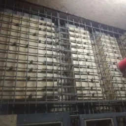 Zbrojenie ściany z siatki stalowej na drewnianym szalunku, widoczny fragment czerwonej rury.