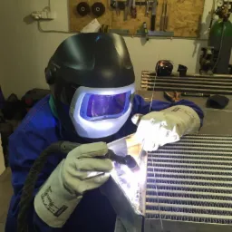 Praca przy użyciu spawarki: spawacz w masce ochronnej łączy metalowe elementy. Widoczne iskry i wyposażenie warsztatowe.