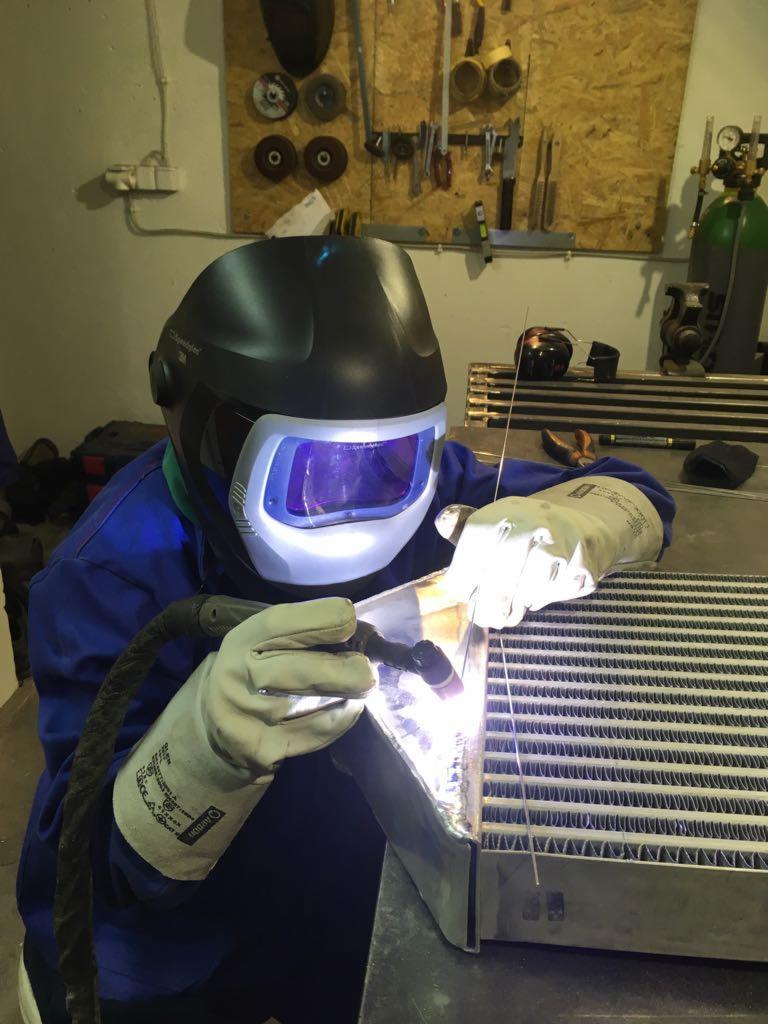 Praca przy użyciu spawarki: spawacz w masce ochronnej łączy metalowe elementy. Widoczne iskry i wyposażenie warsztatowe.