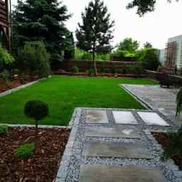 Starannie zaprojektowany ogród z równo przyciętym trawnikiem, ścieżką z płyt betonowych otoczoną drobnym żwirem i korą, oraz ozdobnymi drzewami i krzewami wzdłuż ogrodzenia.