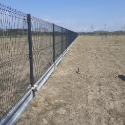 Długi, ciemny panelowy płot z podmurówką, biegnący przez pole w słoneczny dzień, widoczne słupy i siatka ogrodzeniowa, w tle drzewa i niebieskie niebo.