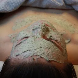 Kobieta leży z zamkniętymi oczami, na twarzy nałożona gruba warstwa maseczki z widocznymi drobinkami alg, głowa spoczywa na szarym ręczniku.