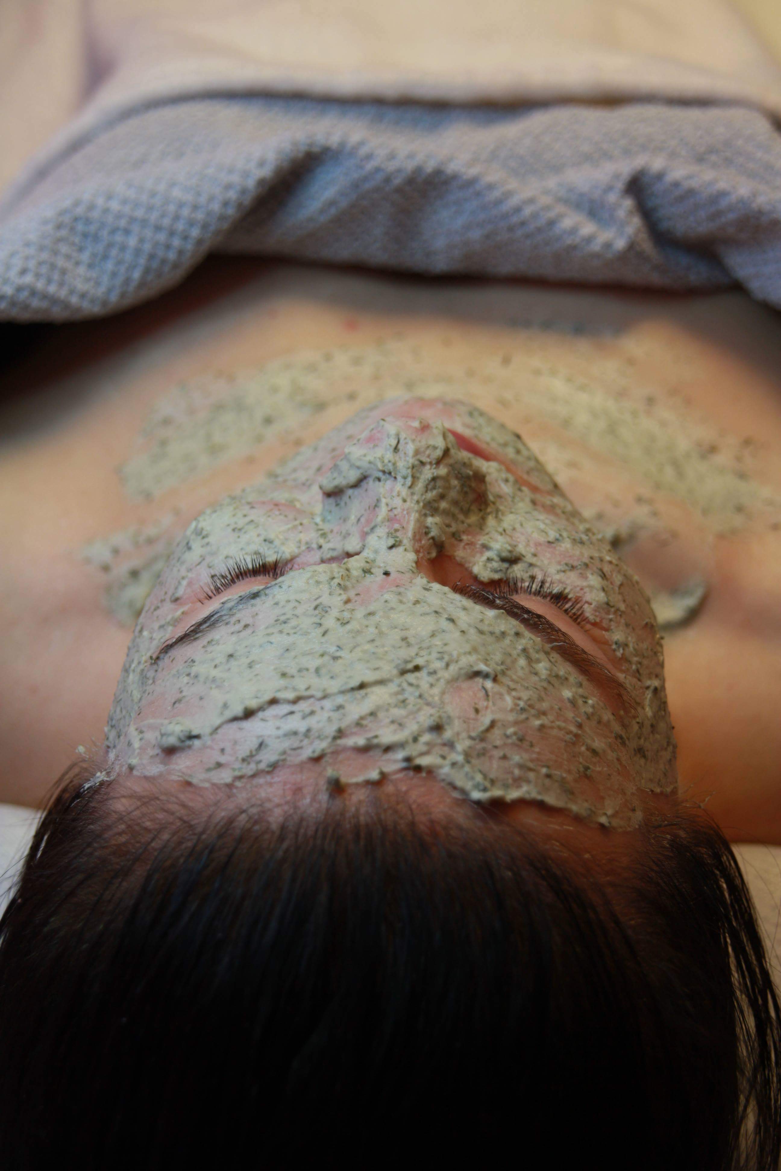 Kobieta leży z zamkniętymi oczami, na twarzy nałożona gruba warstwa maseczki z widocznymi drobinkami alg, głowa spoczywa na szarym ręczniku.