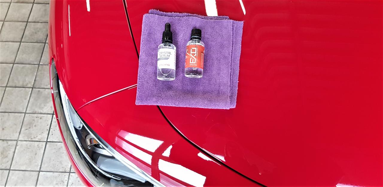 Dwa preparaty do polerowania lakieru samochodowego Crystal Serum Light i EXO na fioletowej ściereczce mikrofibry, leżące na czerwonej karoserii auta, widoczny reflektor