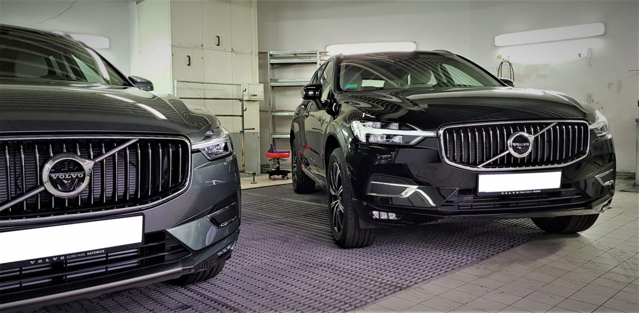 Dwa samochody Volvo SUV, jeden szary i jeden czarny, stojące w dobrze oświetlonym pomieszczeniu z metalową kratą na podłodze, prawdopodobnie w myjni samochodowej.