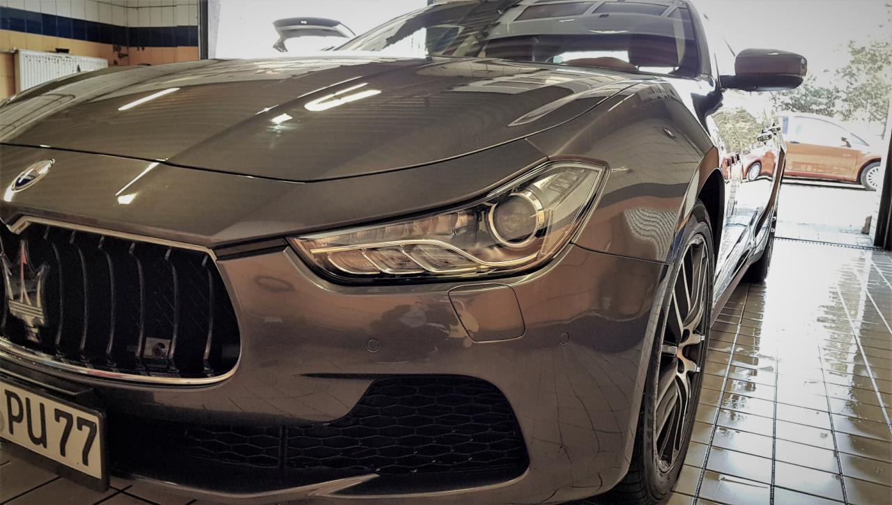 Szary samochód Maserati Ghibli po umyciu, z widocznymi kroplami wody na karoserii i mokrą posadzką myjni, tablica rejestracyjna PU 77, w tle widoczny inny samochód.