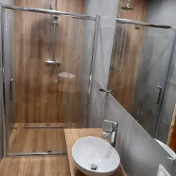 Nowoczesna łazienka z kabiną prysznicową typu walk-in, drewnopodobnymi płytkami i okrągłą umywalką nablatową na drewnianym blacie, odbicie w lustrze.