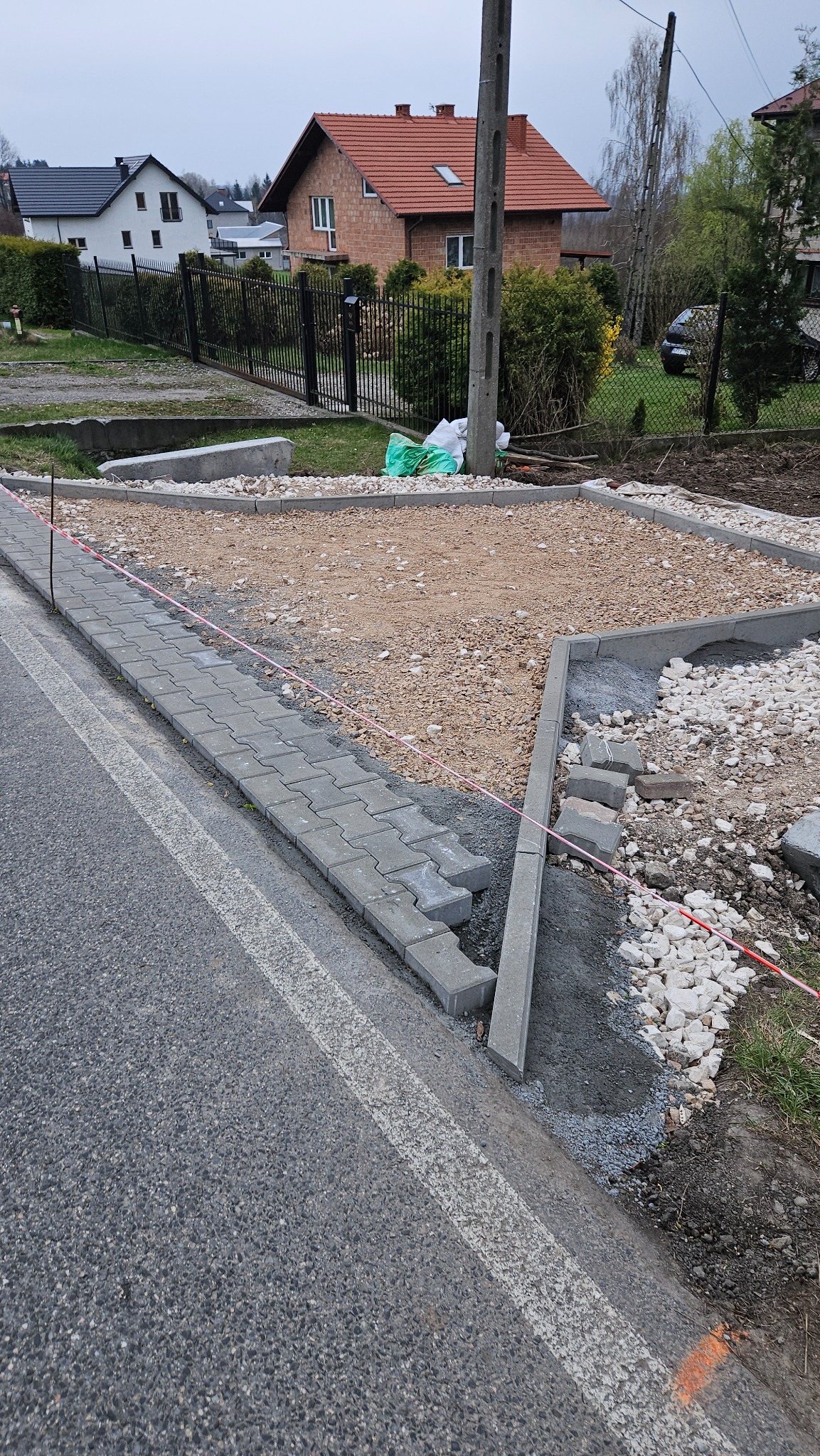 Przygotowanie podłoża pod kostkę brukową przy drodze, widoczne obrzeża, podsypka piaskowa i kamienna, częściowo ułożona kostka wzdłuż krawędzi jezdni, ogrodzenia posesji w tle.