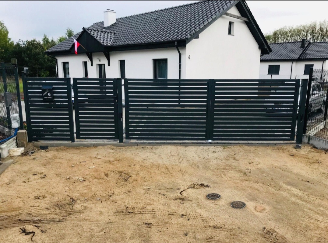 Nowoczesna, pozioma brama przesuwna w kolorze grafitowym, z numerem domu na skrzynce pocztowej, na tle nowego domu z szarą dachówką i polską flagą.