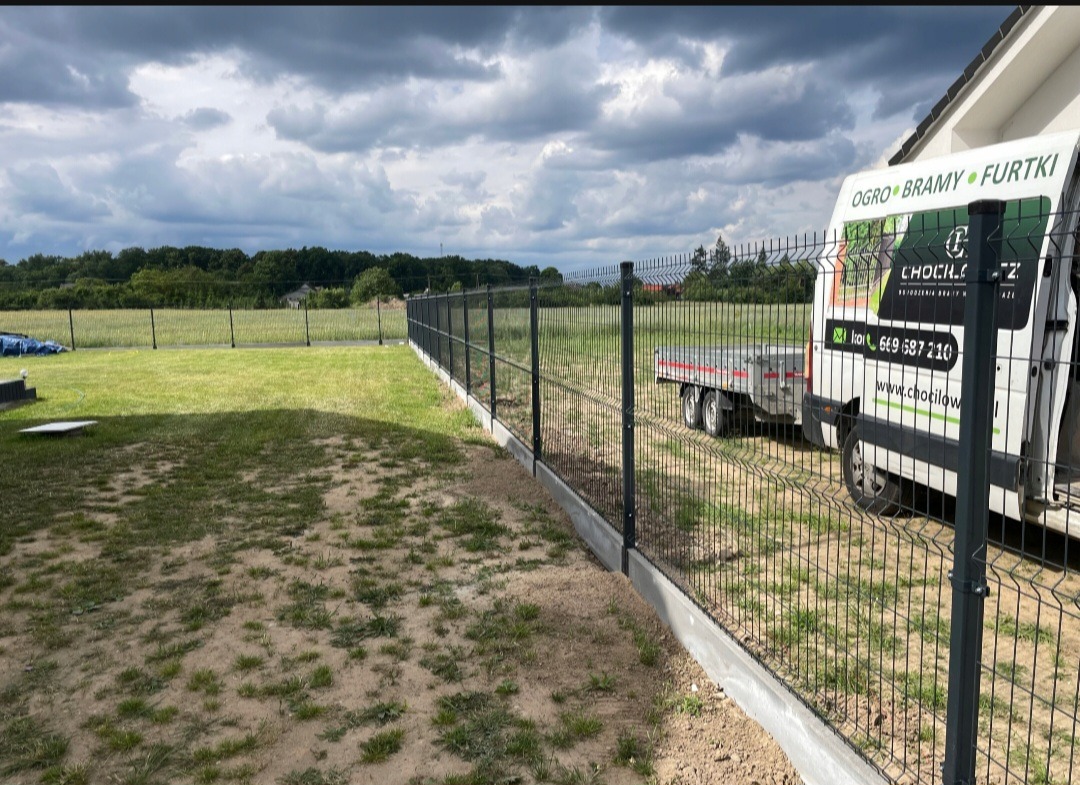 Nowo postawione ogrodzenie panelowe z podmurówką betonową na tle zachmurzonego nieba i zieleni, widoczny samochód dostawczy z logo firmy.