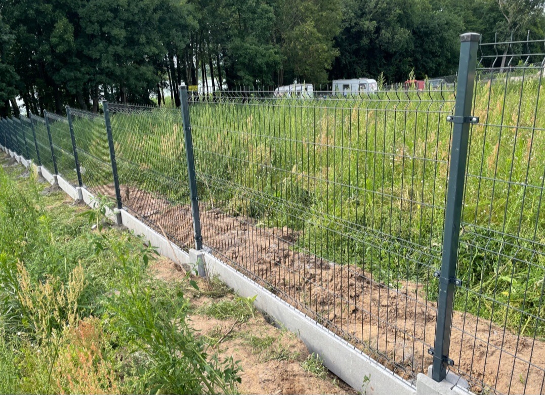 Metalowe ogrodzenie panelowe, świeżo osadzone na betonowej podmurówce, otoczone wysoką trawą i zielenią, z przyczepami kempingowymi w oddali.