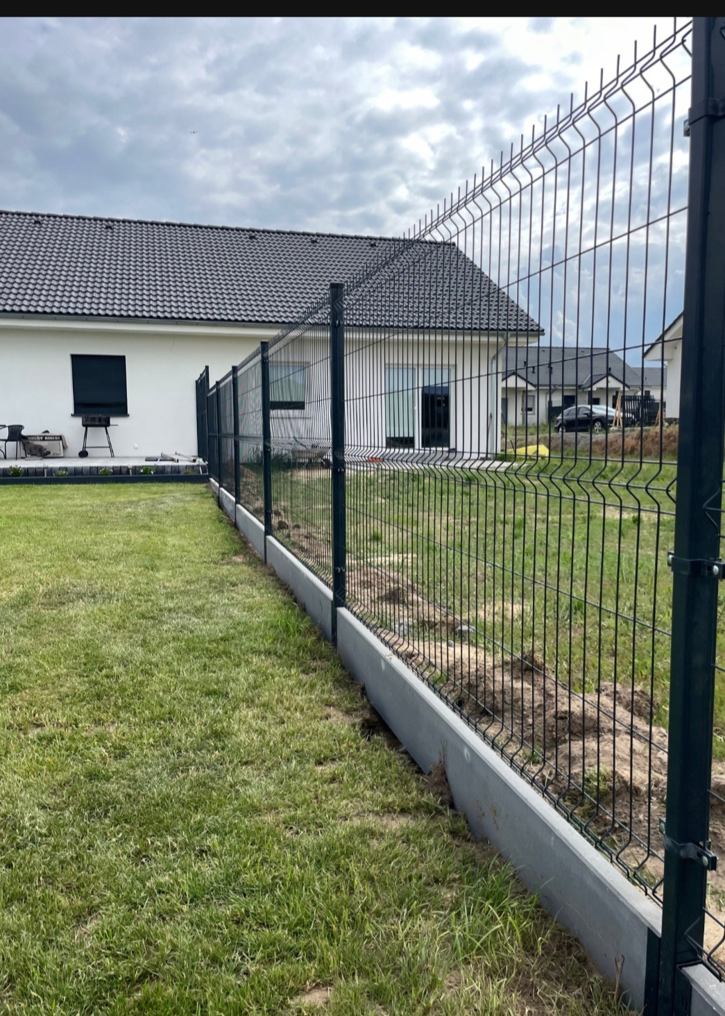 Nowoczesne ogrodzenie panelowe z podmurówką betonową, widok na dom z ciemną dachówką i zielony trawnik.