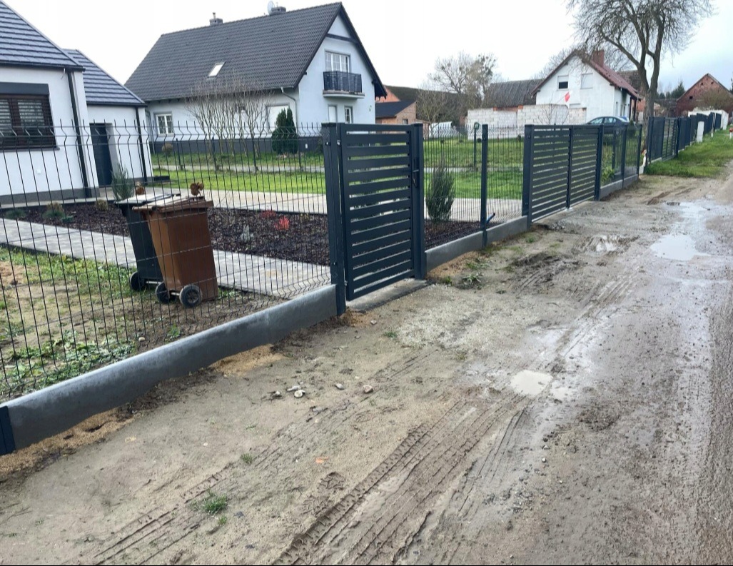 Nowoczesne, grafitowe ogrodzenie panelowe z podmurówką betonową i bramą przesuwną, widoczne z błotnistej drogi w pochmurny dzień, z dwoma pojemnikami na śmieci w tle.