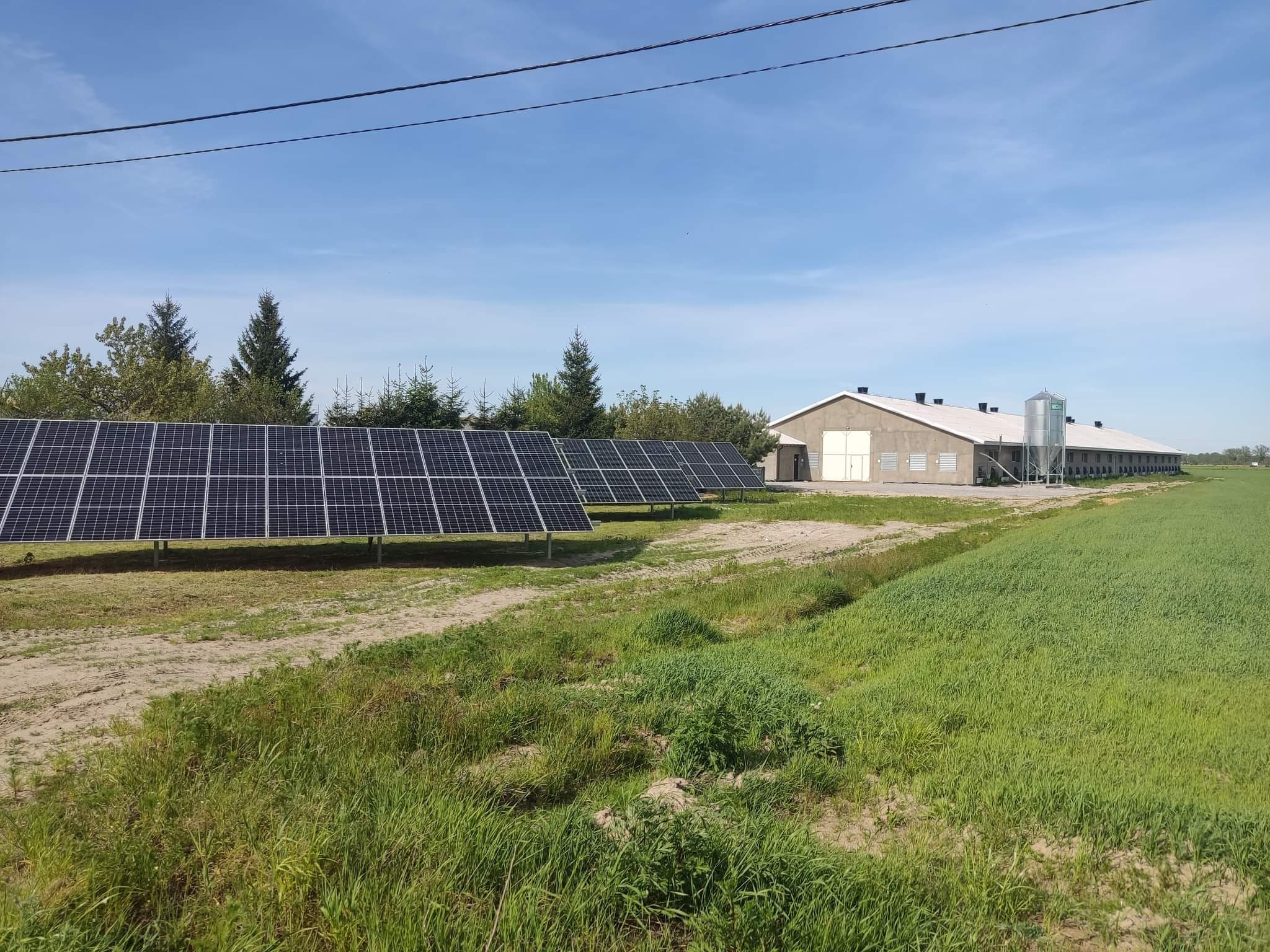 Instalacja paneli słonecznych na trzech stołach, zasilająca gospodarstwo rolne widoczne w tle, w słoneczny dzień.