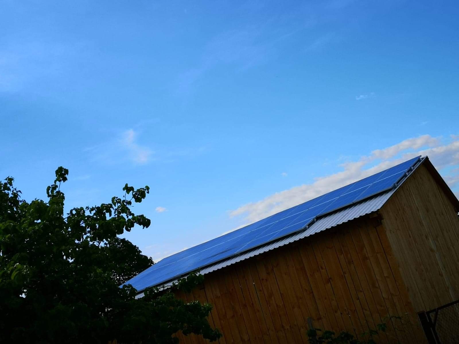 Instalacja fotowoltaiczna 10 kWp na dachu drewnianego budynku gospodarczego, widok z dołu na tle błękitnego nieba z delikatnymi chmurami.