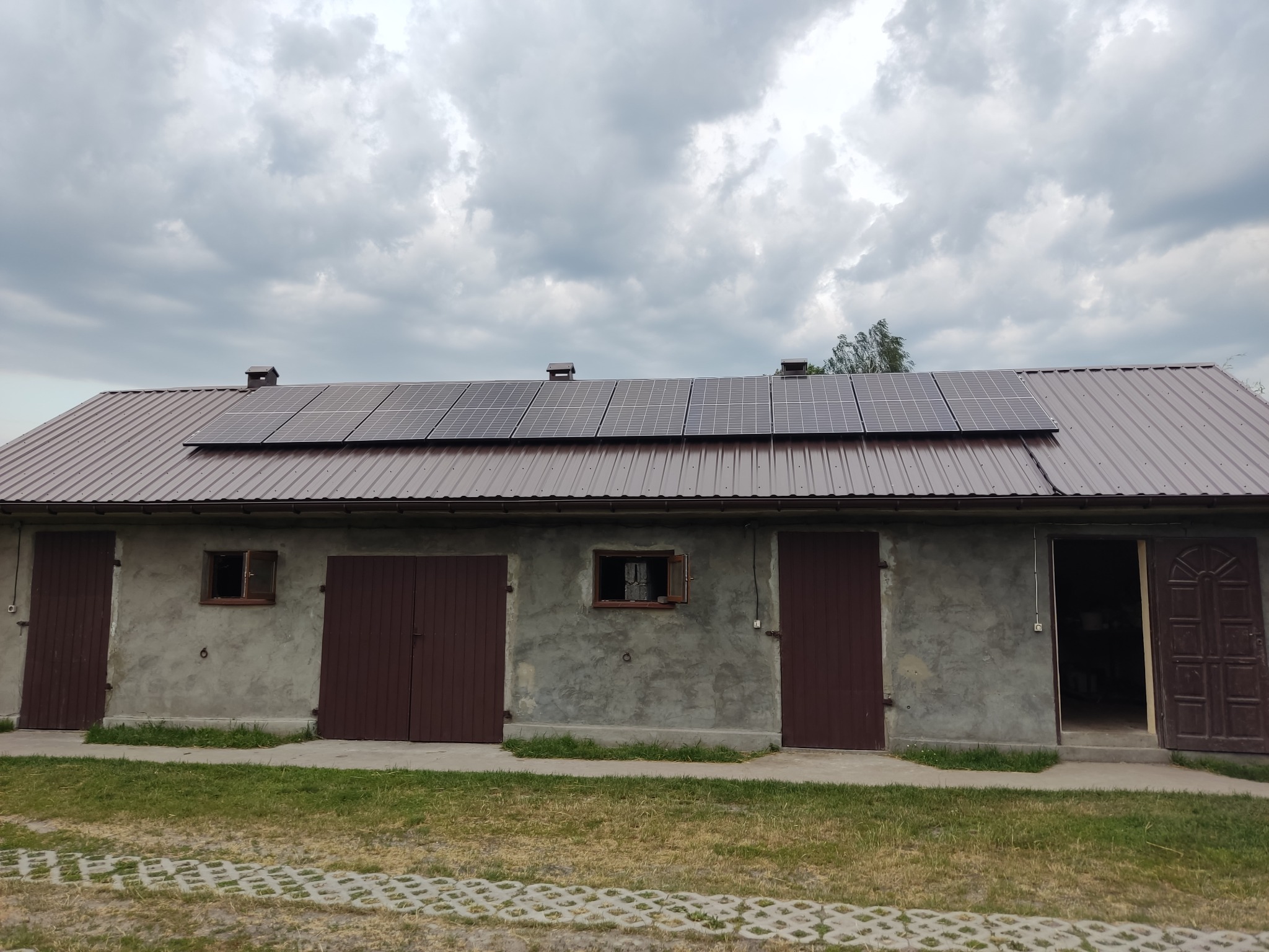 Brązowy budynek gospodarczy z panelami słonecznymi na dachu z blachy trapezowej, widoczne trzy pary brązowych drzwi i dwa małe okna, pochmurne niebo.