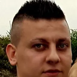 Portret mężczyzny w granatowym garniturze i turkusowej muszce, z krótką fryzurą i wyraźnym spojrzeniem, na rozmytym tle zieleni i fragmentu twarzy dziecka.