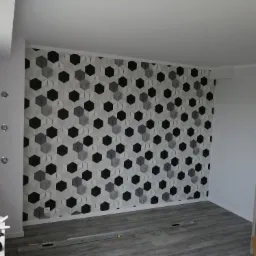 Wykończony pokój z tapetą w geometryczne wzory 3D, szarymi panelami podłogowymi i białymi ścianami, widoczne narzędzia glazurnicze i poziomica na podłodze.