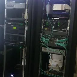 Dwie szafy serwerowe z zainstalowanym sprzętem sieciowym, widoczne kable światłowodowe i zasilające. Górna część szaf oświetlona diodami LED.