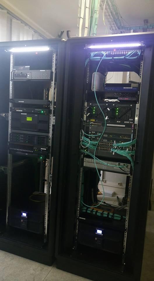 Dwie szafy serwerowe z zainstalowanym sprzętem sieciowym, widoczne kable światłowodowe i zasilające. Górna część szaf oświetlona diodami LED.