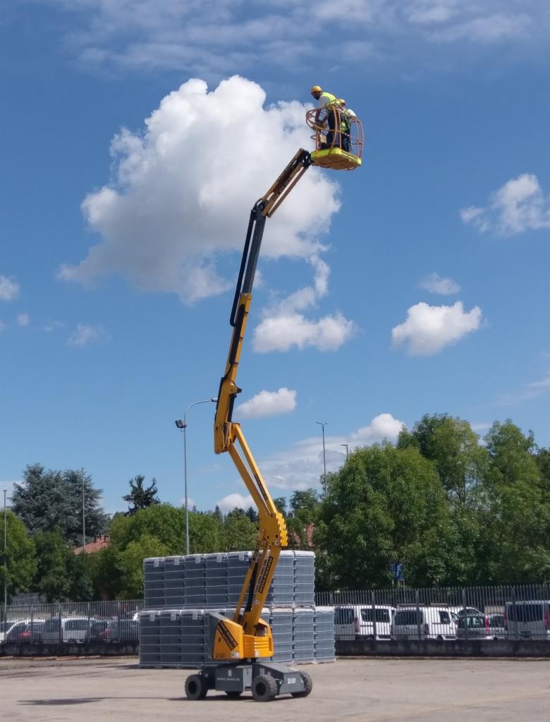 Żółty podnośnik koszowy z pracownikiem w kasku na wysokości, na tle błękitnego nieba z białymi chmurami, zaparkowany na placu obok stosu szarych palet.