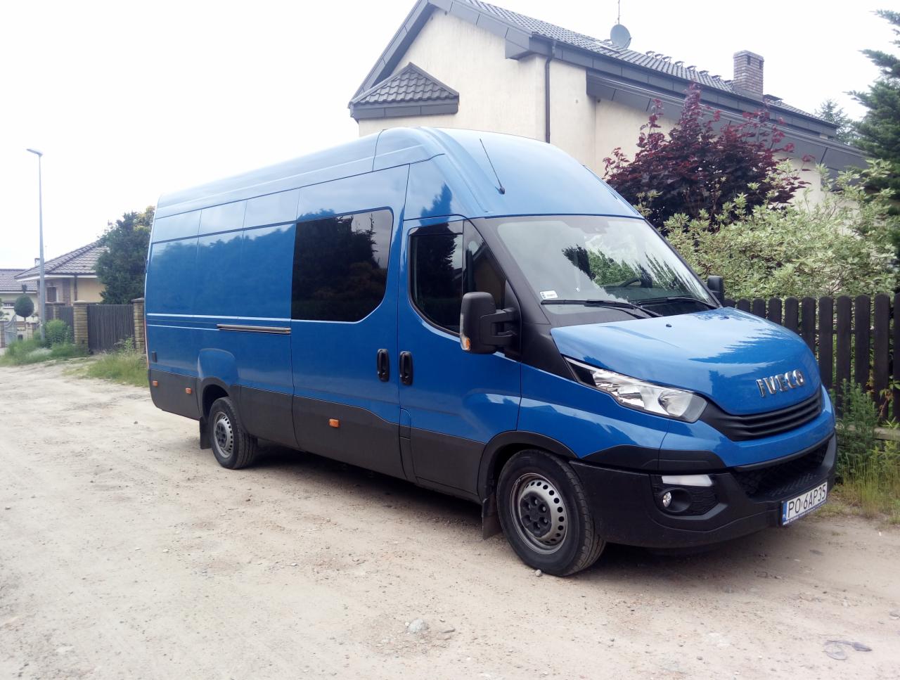 Niebieski furgon dostawczy Iveco z przyciemnionymi szybami, zaparkowany na piaszczystej drodze przed domem jednorodzinnym.