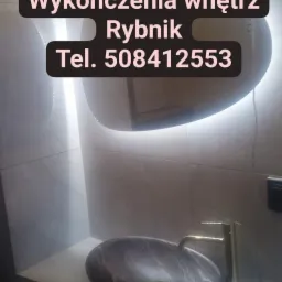 Wykończona łazienka z umywalką z ciemnego marmuru, złotą baterią i podświetlanym lustrem, widoczne płytki na ścianie.