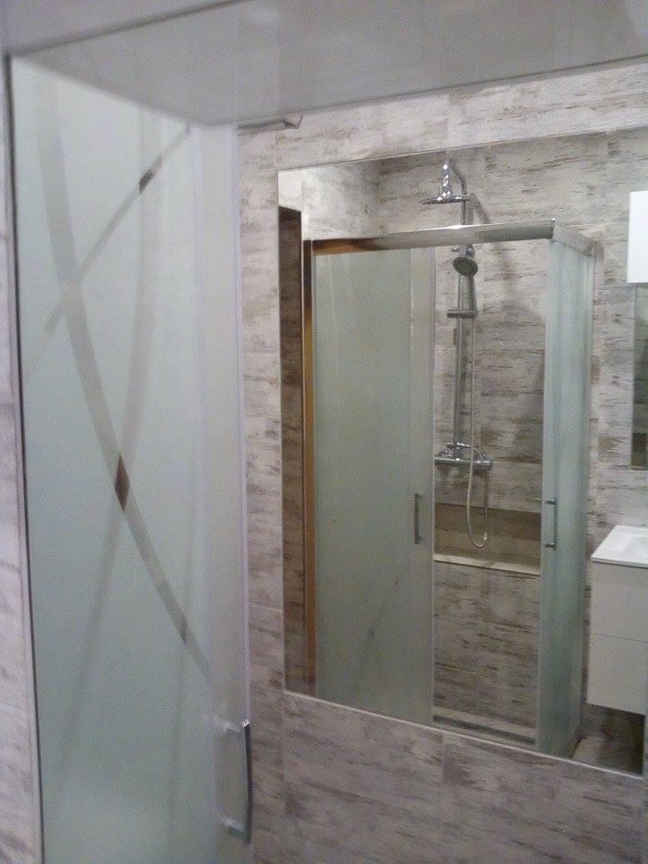 Szklana kabina prysznicowa z matowymi szybami i geometrycznym wzorem, widoczna w lustrze na tle płytek imitujących drewno.