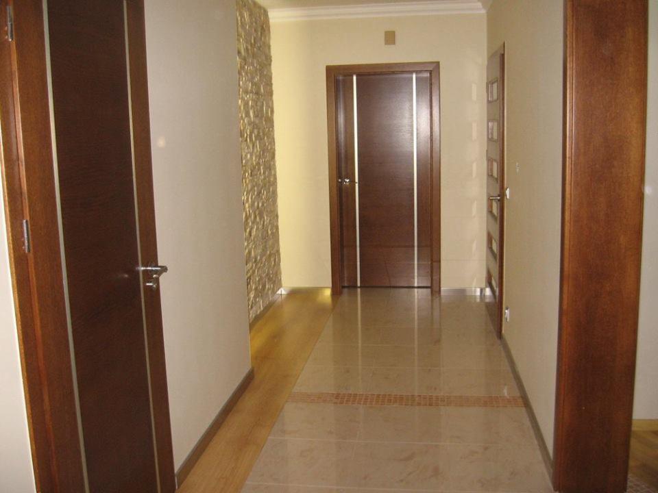 Wnętrze korytarza z brązowymi drzwiami, kamienną ścianą i błyszczącą, beżową podłogą z dekoracyjnym pasem płytek w odcieniach brązu.