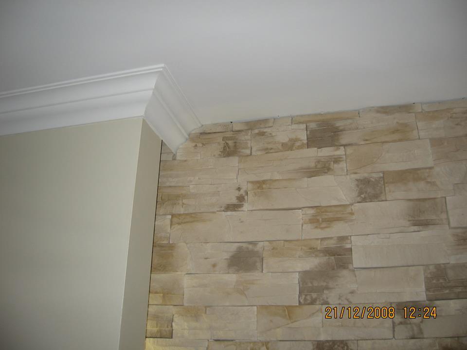 Fragment wnętrza: połączenie gładkiej, beżowej ściany z dekoracyjną ścianą z kamienia w odcieniach beżu i brązu, zakończonej białym, ozdobnym gzymsem pod sufitem.