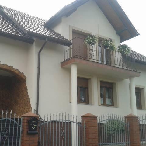 Jasna elewacja dwupiętrowego domu z balkonem ozdobionym kwiatami, widoczna po malowaniu, z brązowymi rynnami i ceglanym wykończeniem wnęki wejściowej oraz metalowym ogrodzeniem z ceglanymi słupkami.