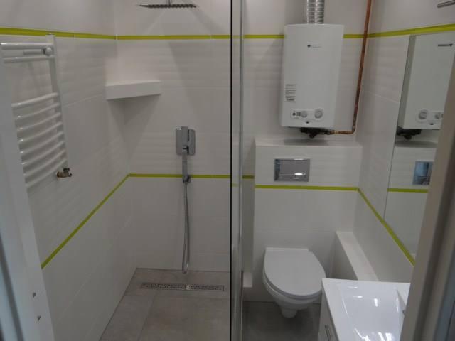 Nowoczesna łazienka z białymi płytkami, kabiną prysznicową typu walk-in, grzejnikiem drabinkowym, podwieszanym WC i piecem gazowym z kominem.