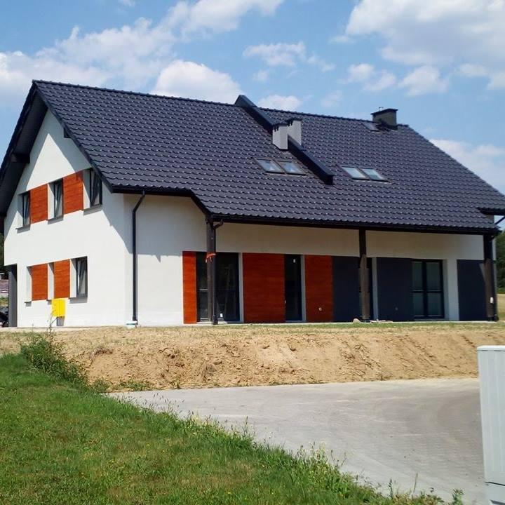 Dwukondygnacyjny dom jednorodzinny z ciemną dachówką i jasną elewacją, drewniane okiennice w kolorze brązowym, widok z zewnątrz, błękitne niebo z chmurami w tle.