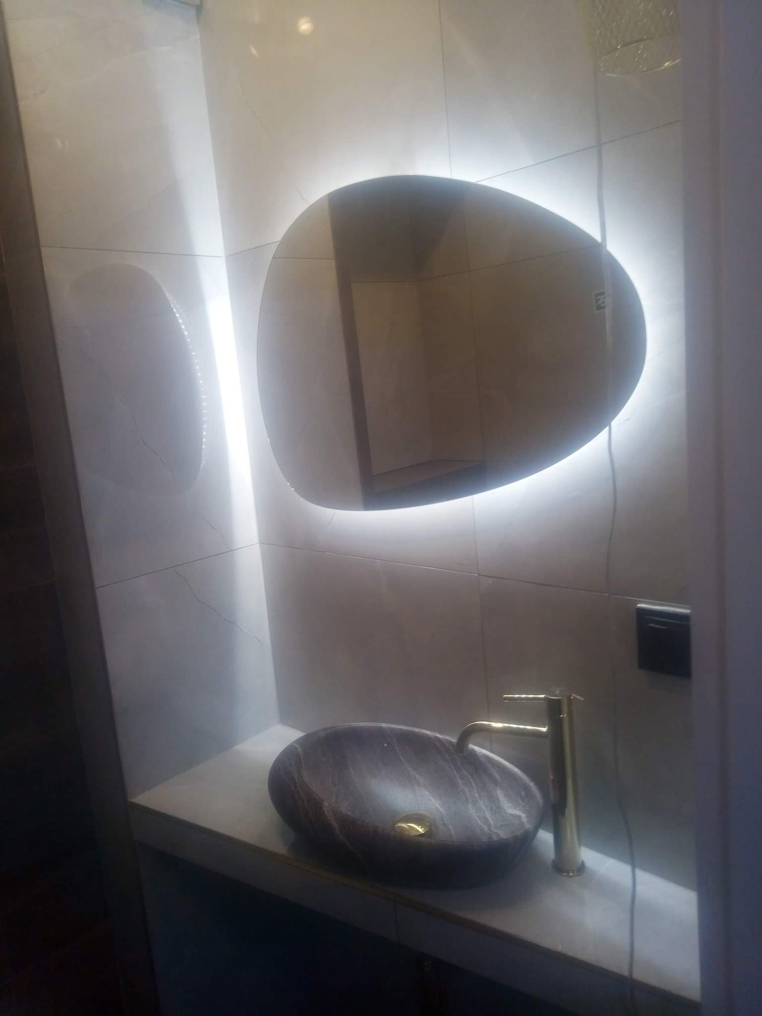 Nowoczesna łazienka z owalnym lustrem podświetlonym LED, marmurowa umywalka nablatowa i złota bateria.