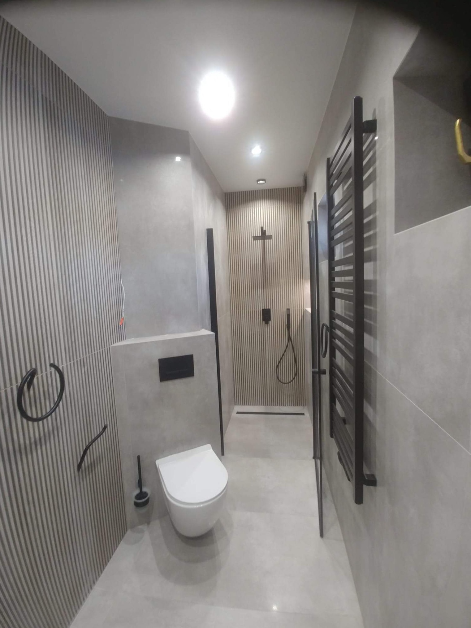 Nowoczesna łazienka z szarymi płytkami imitującymi drewniane lamele, białą toaletą podtynkową, czarnym grzejnikiem drabinkowym i kabiną prysznicową typu walk-in z czarną armaturą.