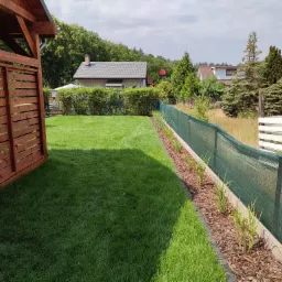 Świeżo założony trawnik z rolki, obrzeża z kory, drewniana altana w rogu, siatka ogrodzeniowa w tle z widokiem na domy i drzewa.