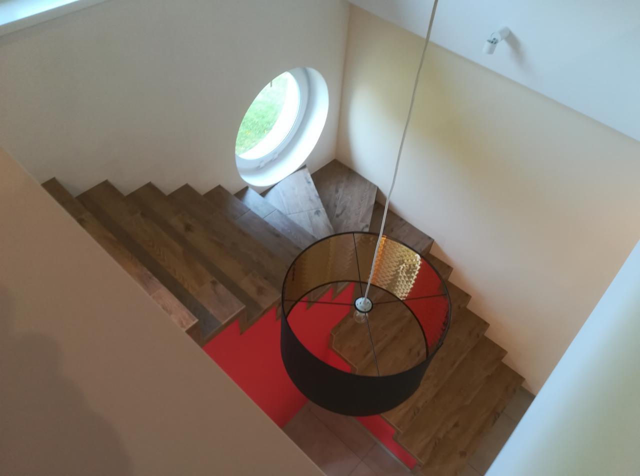 Widok z góry na drewniane schody z okrągłym oknem i nowoczesną lampą z czerwonymi akcentami.