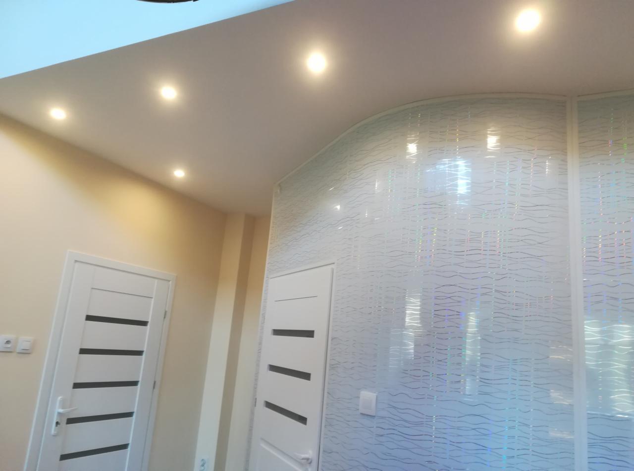 Nowoczesne wnętrze z podwieszanym sufitem i punktowym oświetleniem, dekoracyjna ściana z falistym wzorem, białe drzwi z czarnymi poziomymi paskami.