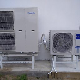 KBI INSTAL Nowoczesne i efektywne instalacje - Dwie jednostki zewnętrzne pomp ciepła, Panasonic z podwójnym wentylatorem i Haier, zamontowane na metalowych stelażach na betonowej podstawie przy białej ścianie budynku.
