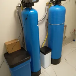 Dwa niebieskie filtry do wody z elektronicznymi panelami sterowania, zainstalowane w pomieszczeniu z białymi płytkami w Wieliszewie. Widoczny także pojemnik na sól i kartonowe pudełko.