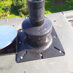 KBI INSTAL Nowoczesne i efektywne instalacje - Czarny kominek wentylacyjny z kratką, zamontowany na szarym dachu z widocznymi śrubami mocującymi i fragmentem metalowego kołnierza uszczelniającego.