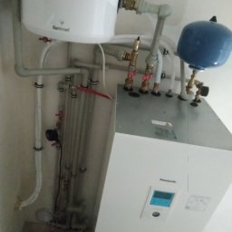 KBI INSTAL Nowoczesne i efektywne instalacje - Biała pompa ciepła Panasonic z podłączonymi rurami i zbiornikiem wyrównawczym w kotłowni, widoczne elementy instalacji hydraulicznej.