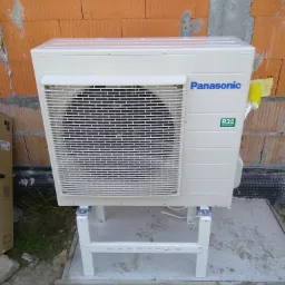 Zewnętrzna jednostka klimatyzacji Panasonic R32 na metalowej podstawie, ustawiona przy ceglanej ścianie w trakcie montażu.