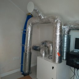 KBI INSTAL Nowoczesne i efektywne instalacje - Biała jednostka centralna rekuperacji z izolowanymi kanałami wentylacyjnymi, aluminiowa folia na rurach, niebieski przewód wentylacyjny przymocowany do ściany, widoczne gniazdka elektryczne.