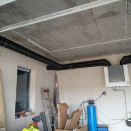 KBI INSTAL Nowoczesne i efektywne instalacje - Instalacja rekuperacji w surowym garażu: czarne rury wentylacyjne pod betonowym sufitem, centrala wentylacyjna na ścianie, niebieski filtr wody, drabina oparta o ścianę, okno z parapetem zagraconym...