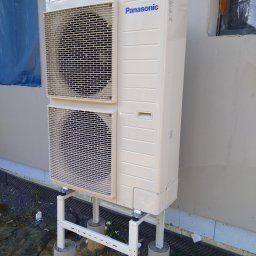 KBI INSTAL Nowoczesne i efektywne instalacje - Beżowa pompa ciepła Panasonic, dwu-wentylatorowa, zamontowana na metalowej konstrukcji osadzonej na betonowych fundamentach z żółtymi śladami, na tle ściany budynku z zabezpieczonym oknem.
